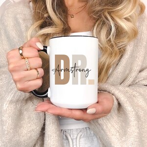 May include: White ceramic mug with a black rim and handle. The mug has the text "DR. Armstrong" printed on it in black and brown.