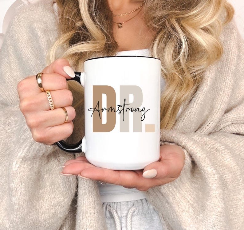 May include: White ceramic mug with a black rim and handle. The mug has the text "DR. Armstrong" printed on it in black and brown.