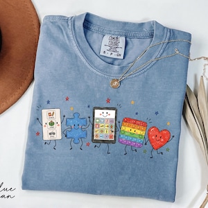 May include: A blue jean colored t-shirt featuring a graphic print of five animated characters: a communication board, a blue puzzle piece, a tablet, a rainbow pop-it toy, and a red heart. The shirt has a Comfort Colors brand tag in the neck.