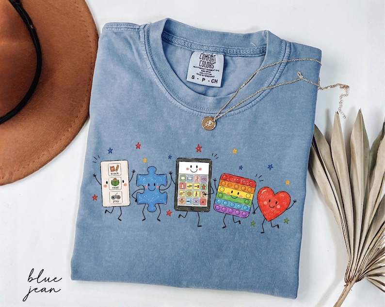 May include: A blue jean colored t-shirt featuring a graphic print of five animated characters: a communication board, a blue puzzle piece, a tablet, a rainbow pop-it toy, and a red heart. The shirt has a Comfort Colors brand tag in the neck.