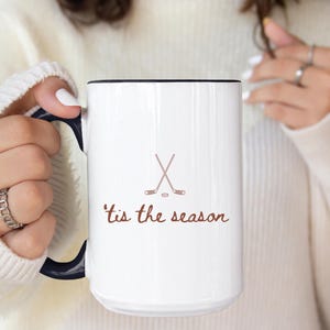 White ceramic hockey mug with crossed sticks and script reading "'tis the season", ideal gift for ice hockey players, fans or coaches in winter.