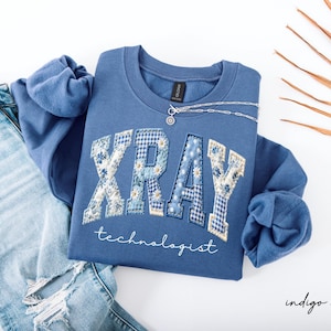May include: Indigo blue sweatshirt with the text "XRAY technologist" in a patchwork design. The letters are decorated with floral and gingham patterns. A silver necklace is draped across the sweatshirt, which is paired with light blue jeans.