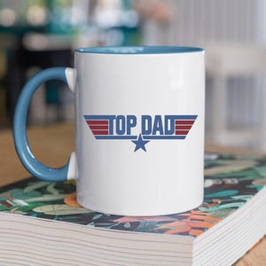 Puede incluir: Taza de cer&aacute;mica blanca con asa e interior azules. La taza presenta el texto "TOP DAD" en un dise&ntilde;o estilizado con detalles en rojo y azul y una estrella azul. La taza est&aacute; sobre un libro con un dise&ntilde;o floral.