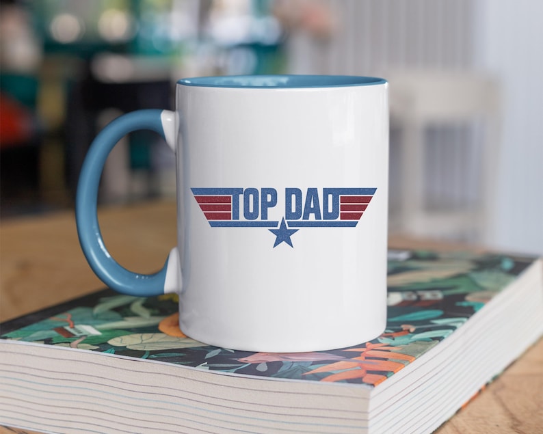 Puede incluir: Taza de cer&aacute;mica blanca con asa e interior azules. La taza presenta el texto "TOP DAD" en un dise&ntilde;o estilizado con detalles en rojo y azul y una estrella azul. La taza est&aacute; sobre un libro con un dise&ntilde;o floral.