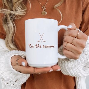 May include: White ceramic mug with a handle, featuring the text "'tis the season" in a brown script, along with crossed hockey sticks and a puck. The mug is held by a person wearing a white sweater and a brown top.