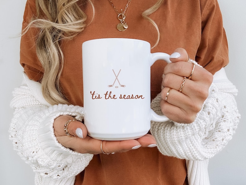 May include: White ceramic mug with a handle, featuring the text "'tis the season" in a brown script, along with crossed hockey sticks and a puck. The mug is held by a person wearing a white sweater and a brown top.