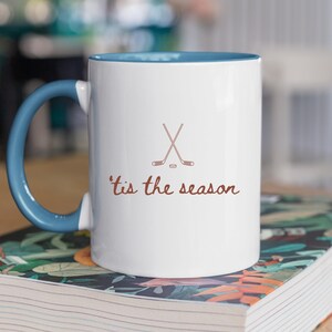 May include: White ceramic mug with a blue interior and handle. The mug features a brown graphic of crossed hockey sticks and a puck, with the phrase "'tis the season" in brown script. The mug is sitting on a book.