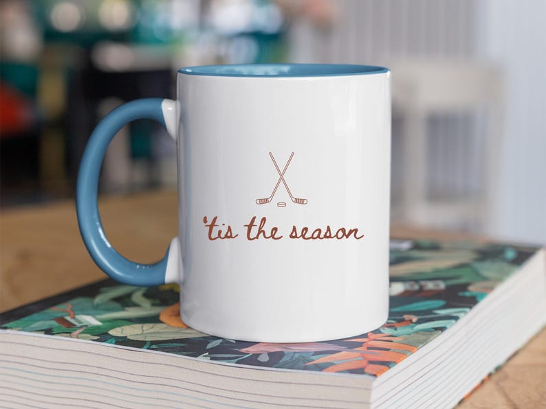 May include: White ceramic mug with a blue interior and handle. The mug features a brown graphic of crossed hockey sticks and a puck, with the phrase "'tis the season" in brown script. The mug is sitting on a book.