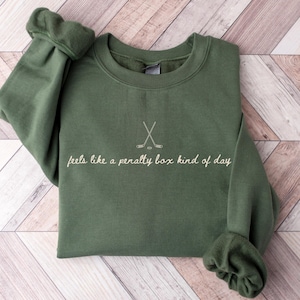 Military Green crewneck sweatshirt with crossed hockey sticks and text "feels like a penalty box kind of day", ideal for hockey players, fans, or coaches.