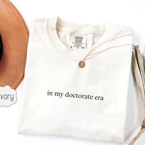 Może przedstawiać: Koszulka w kolorze kości słoniowej z nadrukiem "in my doctorate era" z przodu.