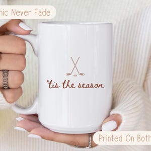 May include: White ceramic mug with a hockey stick and puck design and the words "'tis the season" in brown. The mug has a large handle and is printed on both sides. The text "Graphic Never Fade" and "Printed On Both Sides" are also visible.