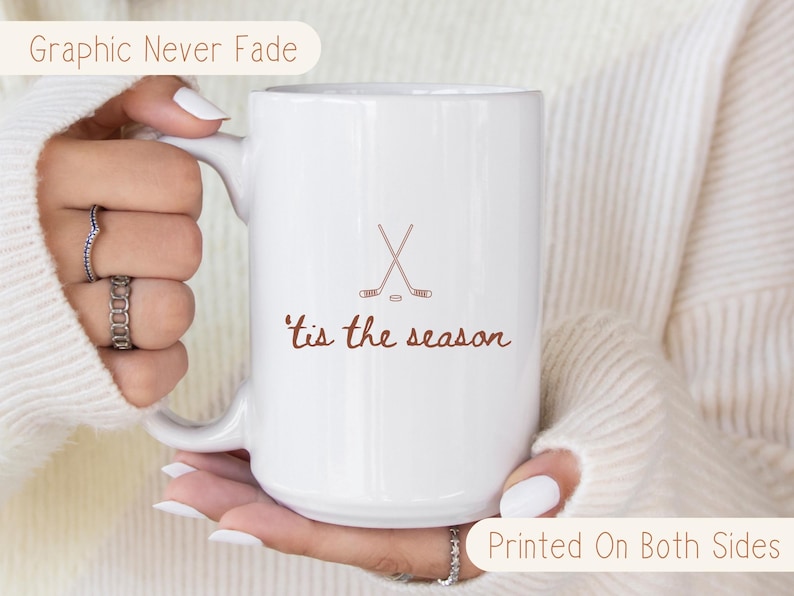 May include: White ceramic mug with a hockey stick and puck design and the words "'tis the season" in brown. The mug has a large handle and is printed on both sides. The text "Graphic Never Fade" and "Printed On Both Sides" are also visible.