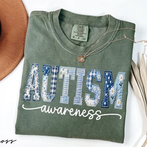 May include: A moss green t-shirt with the word "AUTISM" in blue patchwork letters and the word "awareness" in white script. A gold necklace with a pendant is draped on the shirt.