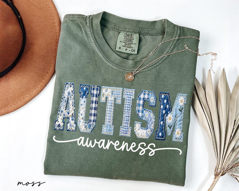 May include: A moss green t-shirt with the word "AUTISM" in blue patchwork letters and the word "awareness" in white script. A gold necklace with a pendant is draped on the shirt.