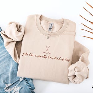 Op de afbeelding: Een zandkleurige sweatshirt met de tekst "feels like a penalty box kind of day" en gekruiste hockeysticks. Een zilveren ketting en een blauwe jeans zijn ook zichtbaar. Het woord "sand" staat rechtsonder.