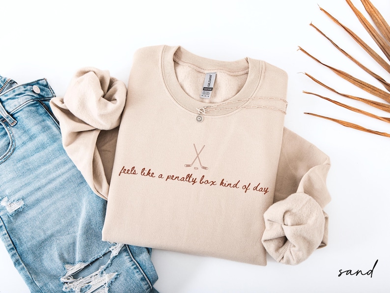 Op de afbeelding: Een zandkleurige sweatshirt met de tekst "feels like a penalty box kind of day" en gekruiste hockeysticks. Een zilveren ketting en een blauwe jeans zijn ook zichtbaar. Het woord "sand" staat rechtsonder.