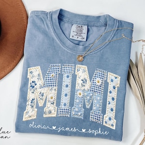 May include: A blue jean Comfort Colors t-shirt with the word "MIMI" appliqued in a patchwork design. The letters feature floral, gingham, and polka dot patterns. The names "olivia", "james", and "sophie" are printed below. A gold necklace and a brown hat are also visible.