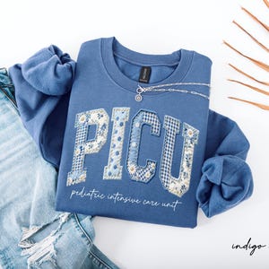 May include: Indigo blue sweatshirt with "PICU" lettering in a patchwork design featuring floral and gingham patterns. The words "pediatric intensive care unit" are below the letters. A silver necklace is draped on the sweatshirt. Light blue jeans are in the background.