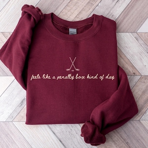 Op de afbeelding: Bordeaux sweatshirt met de tekst "feels like a penalty box kind of day" en een hockeystick-afbeelding. De lange mouwen trui is opgevouwen op een licht houten oppervlak.