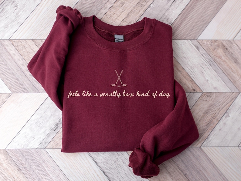 Op de afbeelding: Bordeaux sweatshirt met de tekst "feels like a penalty box kind of day" en een hockeystick-afbeelding. De lange mouwen trui is opgevouwen op een licht houten oppervlak.
