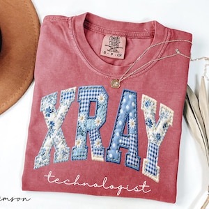 May include: Crimson-colored t-shirt with the word "XRAY" in a patchwork design of blue floral and gingham patterns. The word "technologist" is written below in script. A gold necklace and a hat are also in the image.