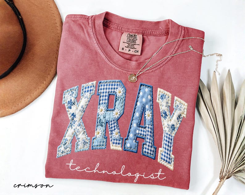 May include: Crimson-colored t-shirt with the word "XRAY" in a patchwork design of blue floral and gingham patterns. The word "technologist" is written below in script. A gold necklace and a hat are also in the image.