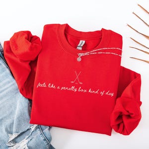 Op de afbeelding: Een felrood sweatshirt met de tekst "feels like a penalty box kind of day" in witte letters, samen met gekruiste hockeysticks. Een zilveren ketting met een hanger ligt op het shirt, naast een spijkerbroek.