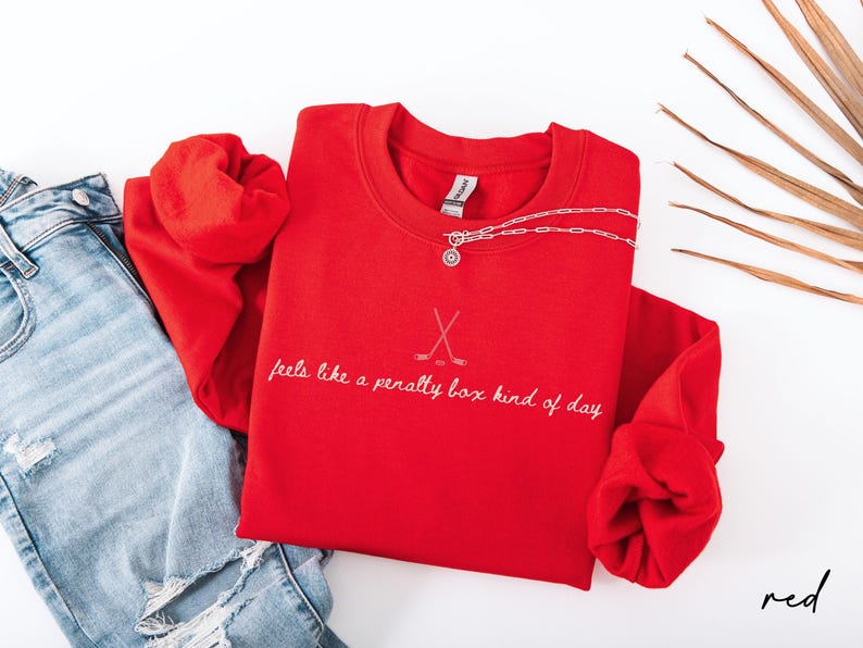 Op de afbeelding: Een felrood sweatshirt met de tekst "feels like a penalty box kind of day" in witte letters, samen met gekruiste hockeysticks. Een zilveren ketting met een hanger ligt op het shirt, naast een spijkerbroek.