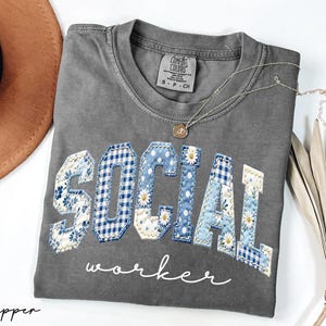 Op de afbeelding: Een grijs Comfort Colors T-shirt met de woorden "SOCIAL WORKER" in een patchwork design. De letters zijn een mix van bloemen-, ruit- en madeliefpatronen in blauw en wit. Een gouden ketting en een bruine hoed zijn ook zichtbaar.