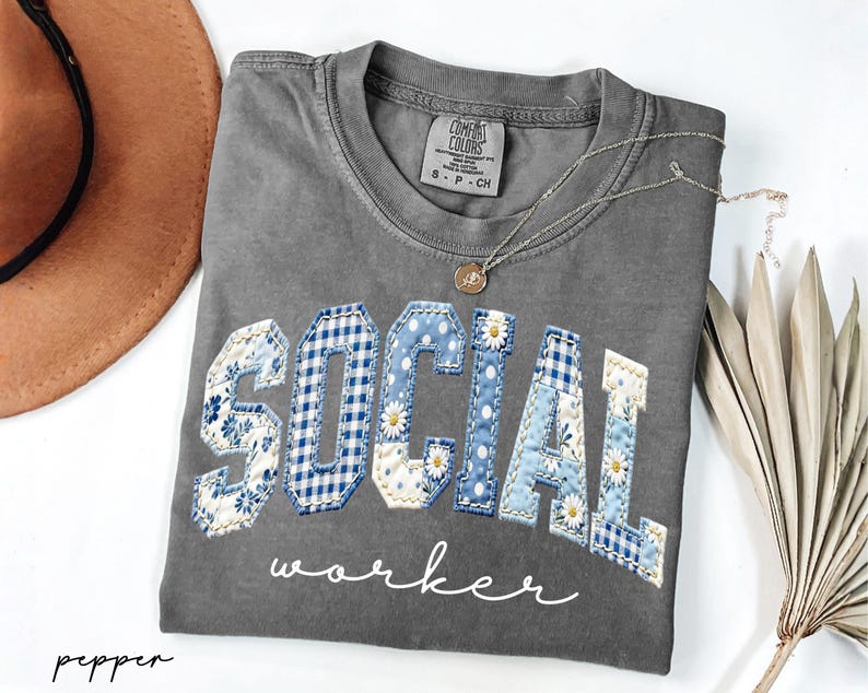 Op de afbeelding: Een grijs Comfort Colors T-shirt met de woorden "SOCIAL WORKER" in een patchwork design. De letters zijn een mix van bloemen-, ruit- en madeliefpatronen in blauw en wit. Een gouden ketting en een bruine hoed zijn ook zichtbaar.