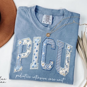 Light blue crewneck shirt with large PICU patchwork letters in floral and gingham fabric, designed for pediatric intensive care nurses, soft cotton relaxed fit.