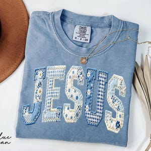 May include: A blue short-sleeve t-shirt with the word JESUS appliqued in a patchwork style using various light blue and white floral and gingham fabrics. The shirt features a crew neckline and a Comfort Colors brand label at the inner collar.