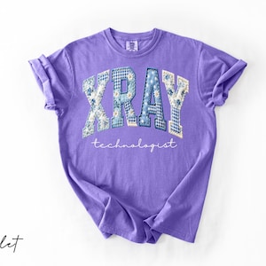 May include: A violet-colored t-shirt with the word "XRAY" in a patchwork design of floral and gingham patterns. The word "technologist" is written in a script font below the word "XRAY".