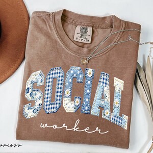 Op de afbeelding: Espresso-kleurig T-shirt met de woorden "SOCIAL WORKER" in een patchwork-ontwerp. De letters zijn een mix van bloemen-, ruit- en stippenpatronen in blauw en wit. Een zilveren ketting met een kleine hanger ligt op het shirt.