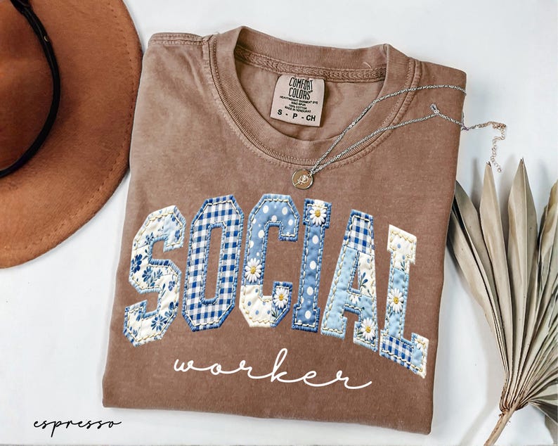 Op de afbeelding: Espresso-kleurig T-shirt met de woorden "SOCIAL WORKER" in een patchwork-ontwerp. De letters zijn een mix van bloemen-, ruit- en stippenpatronen in blauw en wit. Een zilveren ketting met een kleine hanger ligt op het shirt.