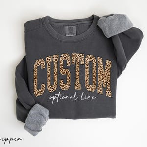 May include: A charcoal gray crewneck sweatshirt featuring the word CUSTOM in a large, block-style font with a brown and black leopard print pattern. Below the word, the text optional line is written in a white, cursive script font.