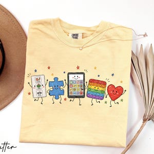 May include: A butter yellow t-shirt featuring a graphic print of five anthropomorphic characters: a white communication board with the text snack, read, play, a blue puzzle piece, a tablet, a rainbow pop-it toy, and a red heart, all with faces.