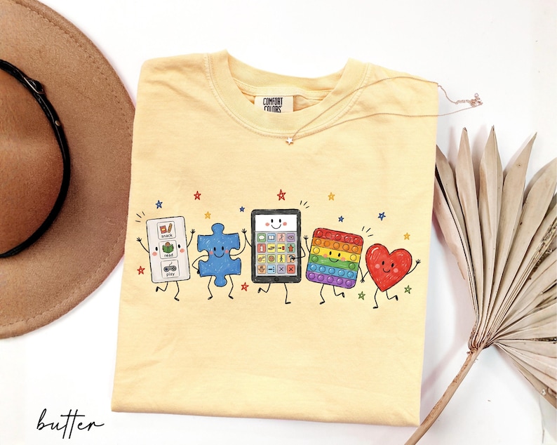 May include: A butter yellow t-shirt featuring a graphic print of five anthropomorphic characters: a white communication board with the text snack, read, play, a blue puzzle piece, a tablet, a rainbow pop-it toy, and a red heart, all with faces.