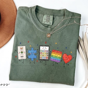 May include: A moss green short-sleeve t-shirt featuring a graphic print of five animated characters: a white communication board with the text snack, read, play, a blue puzzle piece, a tablet, a rainbow pop-it toy, and a red heart, all with faces.