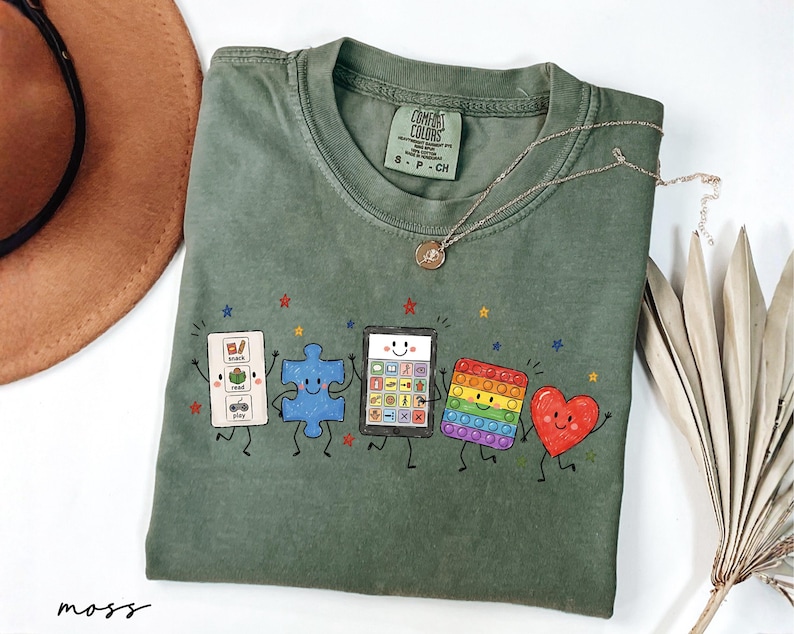 May include: A moss green short-sleeve t-shirt featuring a graphic print of five animated characters: a white communication board with the text snack, read, play, a blue puzzle piece, a tablet, a rainbow pop-it toy, and a red heart, all with faces.
