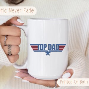 Puede incluir: Taza de cer&aacute;mica blanca con el texto "TOP DAD" en azul y rojo, con un gr&aacute;fico de estrella. La taza tiene un asa grande y el texto "Graphic Never Fade" y "Printed On Both Sides".