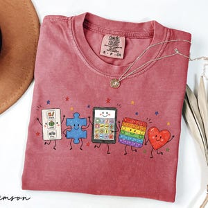 May include: A crimson short-sleeve t-shirt featuring a graphic of five animated characters: a white communication board, a blue puzzle piece, a tablet, a rainbow pop-it toy, and a red heart, all with smiling faces and thin black limbs.