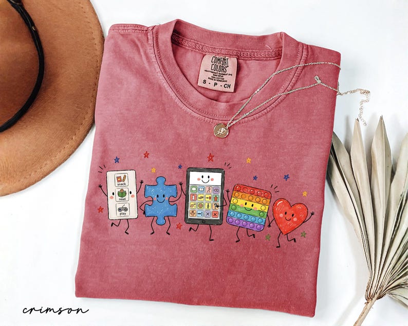 May include: A crimson short-sleeve t-shirt featuring a graphic of five animated characters: a white communication board, a blue puzzle piece, a tablet, a rainbow pop-it toy, and a red heart, all with smiling faces and thin black limbs.