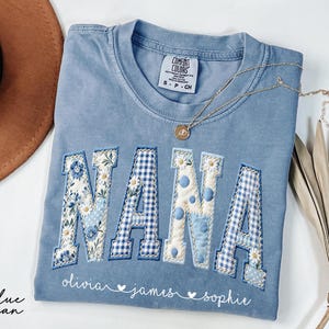 May include: A blue jean Comfort Colors t-shirt with the word "NANA" appliqued in floral, polka dot, and gingham patterns. The names "olivia", "james", and "sophie" are embroidered below. A gold necklace and a brown hat are also visible.