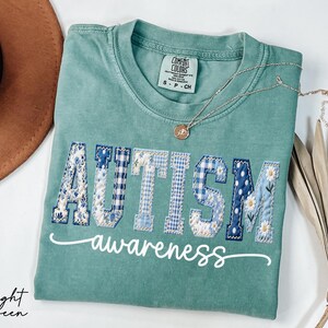May include: Light green t-shirt with the word "AUTISM" in patchwork letters, featuring floral, gingham, and polka dot patterns. The word "awareness" is written in cursive below. A gold necklace and a hat are also visible.