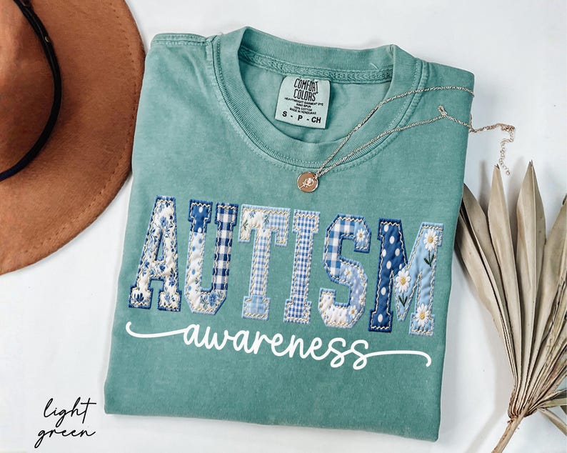 May include: Light green t-shirt with the word "AUTISM" in patchwork letters, featuring floral, gingham, and polka dot patterns. The word "awareness" is written in cursive below. A gold necklace and a hat are also visible.