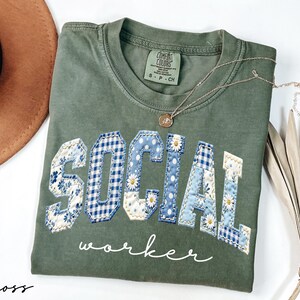 Op de afbeelding: Een mosgroen t-shirt met de woorden "SOCIAL WORKER" in een patchwork-ontwerp. De letters zijn een mix van bloemen- en ruitpatronen in blauw en wit. Een gouden ketting met een kleine hanger ligt op het shirt.
