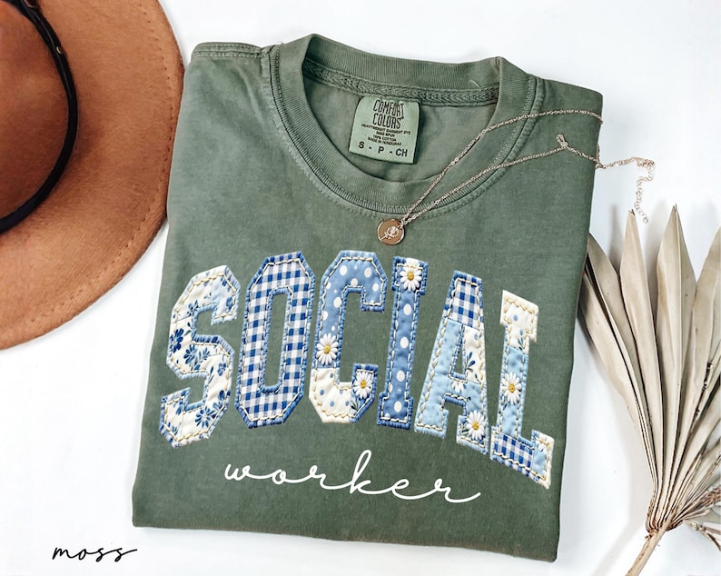 Op de afbeelding: Een mosgroen t-shirt met de woorden "SOCIAL WORKER" in een patchwork-ontwerp. De letters zijn een mix van bloemen- en ruitpatronen in blauw en wit. Een gouden ketting met een kleine hanger ligt op het shirt.
