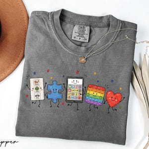 May include: A charcoal gray short-sleeve t-shirt featuring a graphic print of five animated characters: a white communication board, a blue puzzle piece, a tablet, a rainbow-colored pop-it toy, and a red heart, all with arms and legs.