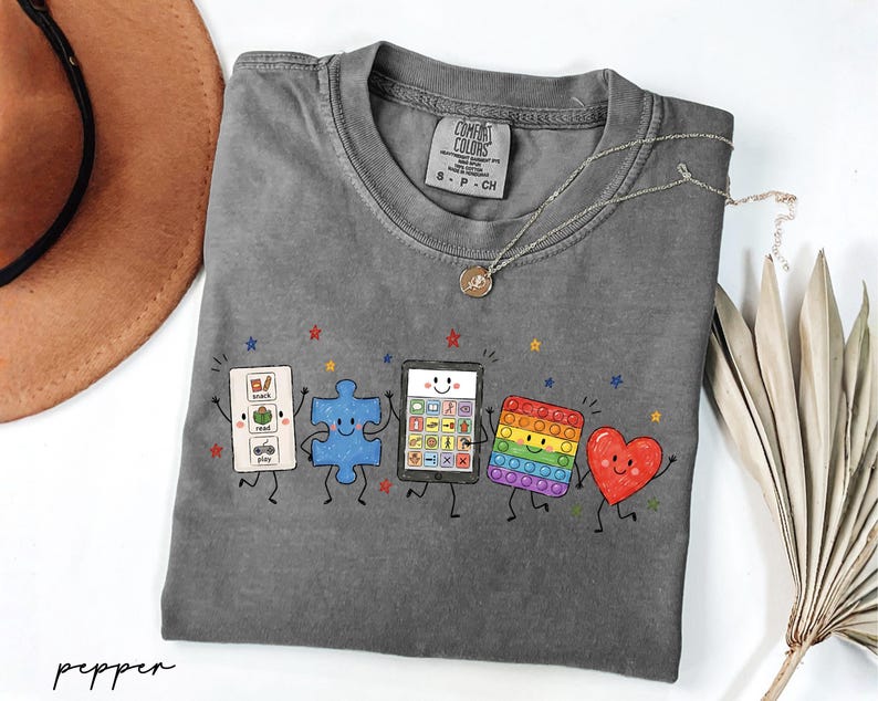 May include: A charcoal gray short-sleeve t-shirt featuring a graphic print of five animated characters: a white communication board, a blue puzzle piece, a tablet, a rainbow-colored pop-it toy, and a red heart, all with arms and legs.